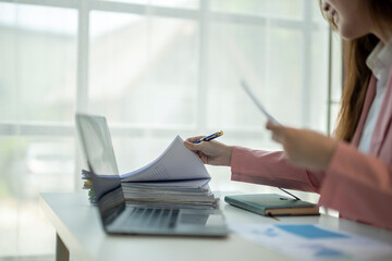 A woman is sitting at a desk with a laptop and a stack of papers. She is writing on the papers with a pen