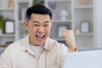 Joyful middle-aged Asian man in a casual shirt celebrating a triumph in front of his laptop, expressing excitement and happiness in a home office setting.
