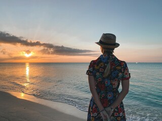 Woman in hat and cute summer dress standing at the beach, looking at sunrise