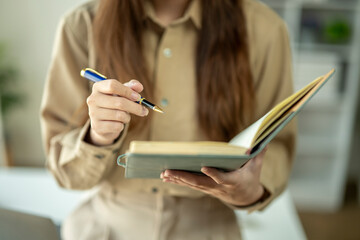 A woman is holding a pen and looking at a book. She is likely writing or taking notes from the...