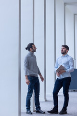 deux jeune hommes d'affaire ou employés de bureau discutent debout dans les locaux de leur entreprise. l'un d'eux porte un ordinateur portable