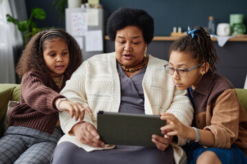 Front view portrait of Black senior woman using digital tablet at home with two children helping - Powered by Adobe