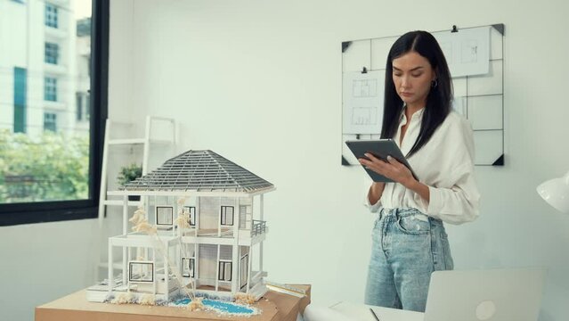Beautiful caucasian architect focus on using tablet to check house model on meeting table with blueprint, house model and architectural document scatter around at meeting room. Immaculate.