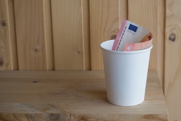 A white paper cup with a euro bill inside and the Eurozone symbol on the bill on a wooden table,...