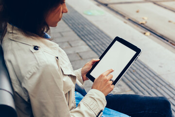 Side view of stylish dressed young woman touching with finger on mock up screen of modern touch pad connected to 4G internet doing shopping online while waiting on transport sitting on tram stop