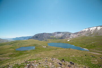 Two lakes, sunlit green meadow, alpine valley and mountain range. Blue sky gradient reflected in water. Top view from rocks to dirt road between two beautiful lakes in high mountains in sunny day.