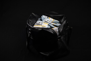 On a black background there is a bag with stacks of hundred dollar bills. A black leather bag full...