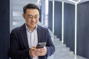 A cheerful Asian businessman in a suit stands using his mobile phone in a contemporary office...
