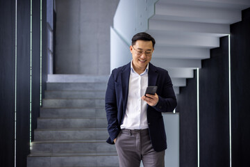 A cheerful Asian businessman in a suit and glasses, using a smartphone while descending a stylish staircase in a contemporary office setting.
