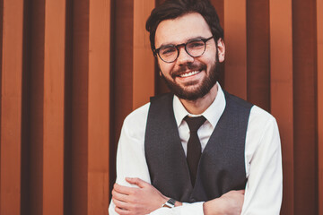 Half length portrait of happy positive hipster guy standing near wall in trendy waistcoat smiling and looking at camera, successful cheerful man in stylish spectacles posing on publicity area