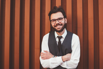 Half length portrait of happy positive hipster guy standing near wall in trendy waistcoat smiling and looking at camera, successful cheerful man in stylish spectacles posing on publicity area