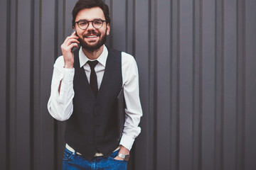 Half length portrait of cheerful hipster guy looking at camera during positive international telephone conversation outdoors, positive man in spectacles using roaming connection on publicity area