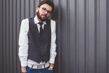 Uncertain caucasian hipster guy in spectacles standing near promotional background and looking at camera outdoors, young male student in casual wear posing near copy space area for advertising text