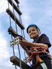 A teenage boy in sports safety gear climbs a hanging rope ladder in a rope amusement park. View...