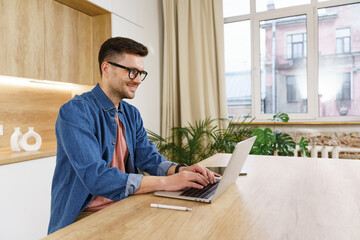 A content young man in glasses types on a laptop in a naturally lit office with a view of the city,...