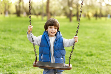 Outdoors portrait of cute preschool laughing boy swinging on a swing at the playground in nature park, summer sunny day