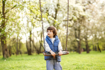 Outdoors portrait of cute preschool laughing boy swinging on a swing at the playground in nature park, summer sunny day