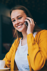 Portrait of cheerful hipster girl laughing at camera during smartphone conversation in cafe interior, happy woman casual dressed enjoying friendly telephone call in cafeteria during tea time