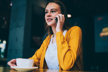 Positive charming hipster girl talking during telephone conversation on cellular sitting in cafeteria, beautiful woman calling on smartphone using good high speed connection enjoying coffee break