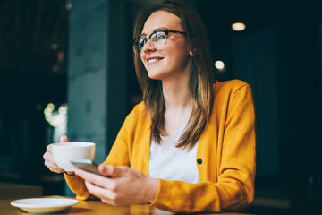 Happy woman in spectacles for vision correction holding smartphone gadget and cup with tea in hands, positive hipster girl looking away while using good mobile connection for communicate in cafeteria