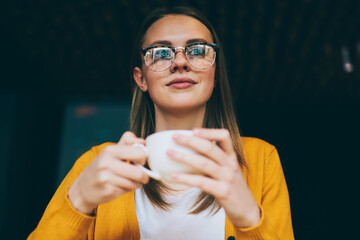 Positive female teenager enjoying tea time in cafeteria while dreaming about upcoming trip with friends, charming hipster girl in stylish eyewear holding cup with caffeine beverage and looking away