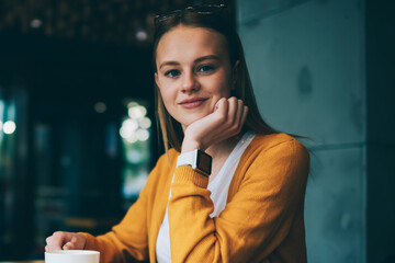 Charming caucasian hipster girl casual dressed smiling and looking at camera while holding cup with hot tea, positive woman enjoying free time on leisure feeling happy of resting in cafeteria