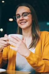 Portrait of positive woman in stylish spectacles for provide eyes protection laughing and looking at camera during coffee break in cafeteria, successful hipster girl holding cup with caffeine beverage