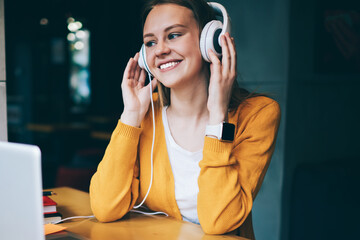 Cheerful hipster girl listening positive playlist via multimedia application and earphones, happy...