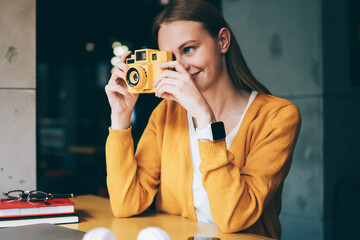 Positive hipster girl casual dressed focusing on vintage camera and making photos indoors, successful amateur photographer taking pictures via old fashioned equipment enjoying hoppy in cafeteria - Powered by Adobe