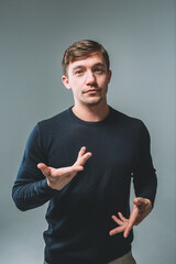 A young man in a blue sweater on a gray background. The man spreads his hands, expressing doubts...