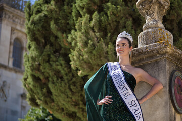Young, pretty, blonde woman in green party dress with sequins, diamond crown and beauty pageant winner's sash, staring into infinity proudly leaning on a column. Concept of beauty, fashion, pageant. - obrazy, fototapety, plakaty