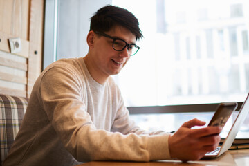 Positive student in optical spectacles reading good news on modern mobile phone while working at laptop computer in stylish coworking space.Graphic designer in eyeglasses chatting on cellular