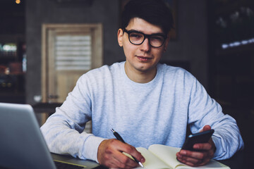Portrait of handsome young man in eyeglasses holding mobile phone in hand and making notes in notepad sitting at laptop computer device.Male administrator looking at camera while writing text