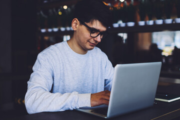 Young handsome student in optical spectacles chatting online with friends in social networks using laptop device.Male designer typing text on keyboard of computer with high speed internet connection