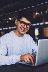 Portrait of successful it professional in stylish eyeglasses working at modern laptop computer and using 4G high speed internet connection while looking at camera and smiling sitting in coworking