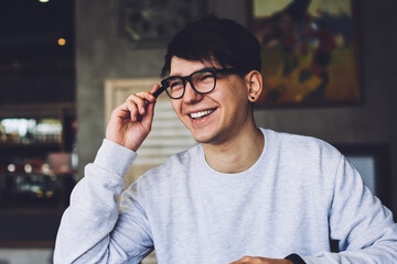 Positive young man in spectacles with good lenses remembered funny case of life for writing interesting story sitting at coffee table in stylish cafeteria.Cheerful student laughing during homework