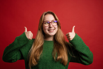 portrait of a happy girl in glasses holding thumbs up on red background