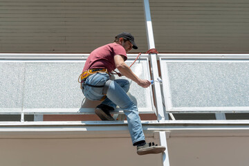 A man in high-altitude climbing equipment paints with a roller at height on the balcony of the house, dangerous work.