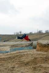 A guy in a red sweater does an acrobatic back somersault from a small ledge on the seashore in autumn
