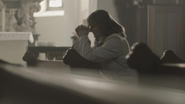 Christian female parishioner sitting on pew bench, praying to God with clenched fists side view medium shot. Mid adult caucasian woman during prayer in empty church