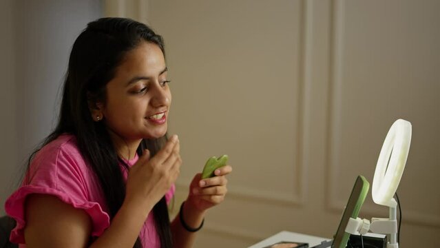 On the side a happy brunette Indian girl does a facial massage using a special massager and talks about it during her Beauty blog about beauty and make-up in a modern apartment during the day in front