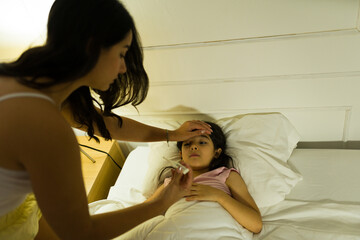 Latina mother gently checks her daughter's forehead for fever in the comforting glow of the bedroom night