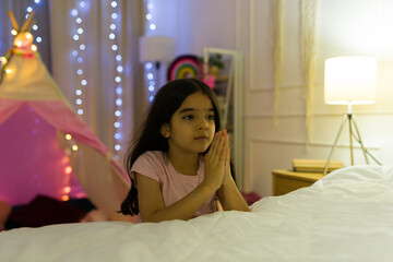 Hopeful young latina girl prays in her warmly lit bedroom at home during the nighttime