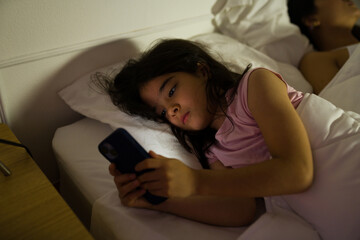 Closeup of a little girl using a smartphone at night while her mother sleeps beside her