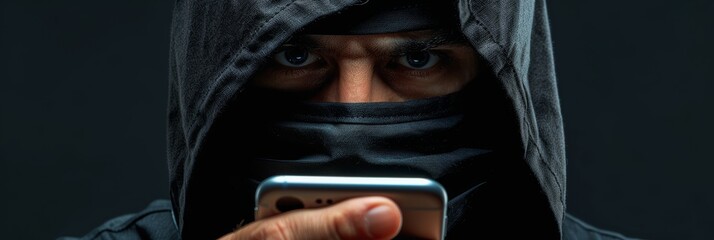 Male cybercriminal in balaclava and black attire holding phone, illustrating phone scam scheme