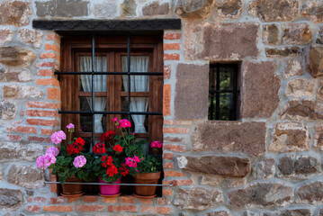 A window with a flower pot on the sill on a stone building wall. The window has bars and pink flowers