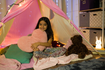Happy young latina girl has fun in her bedroom, cuddling with a heart pillow in an improvised tent