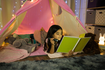 Cute little girl immersed in a book inside a homemade tent in her bedroom, creating a magical nighttime atmosphere