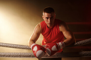 Young serene man in red vest and boxing gloves standing on ring and looking aside while having...