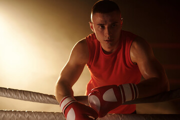Tired and sweaty boxer in red sportswear and boxing gloves looking aside while standing in boxing...